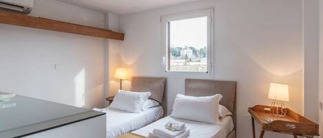 Camera d'albergo con due letti, una testiera in legno, una finestra con vista, un comodino con una lampada e un comò in vetro.