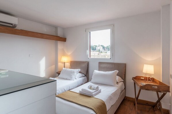 A hotel room with two beds, a wooden headboard, a window with a view, a bedside table with a lamp, and a glass dresser.