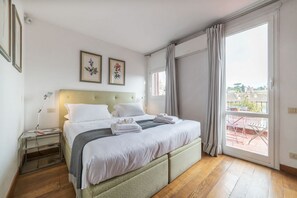 A neatly made bed with white linens and towels, a bedside table with a lamp, and a window with curtains.