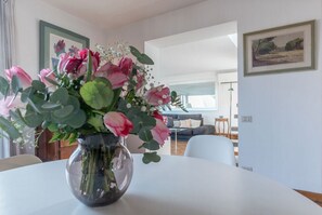 Un comedor con una mesa redonda, un jarrón de flores y vistas a una sala de estar con un sofá y un cuadro en la pared.
