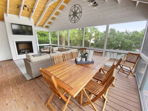 Une terrasse extérieure avec une table et des chaises en bois, une cheminée et une grande fenêtre donnant sur un paysage verdoyant.