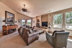 A spacious living room with a leather sofa, a grey armchair, a coffee table, and a television. There is a fireplace, a large window, and a painting on the wall.