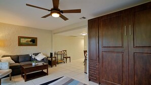 A living room with a sofa, coffee table, and a ceiling fan. There is a wooden wardrobe and a painting on the wall. - Oceanfront complex at Casa De Emdeko unit #130 (Kailua-Kona)