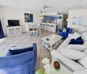 Uma sala de estar moderna com televisão, móveis brancos, um espaço de jantar e uma cozinha ao fundo.