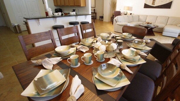 A dining table set with plates, bowls, and cups, surrounded by chairs in a room with a kitchen and a living area. - Ly231617 - Watersong - 6 Bed 4 Baths Villa (Davenport)