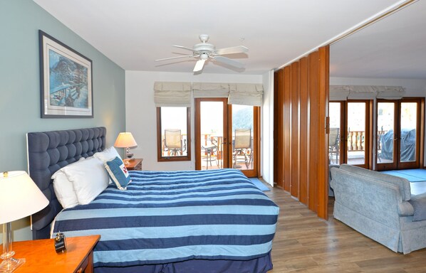 A bedroom with a large bed, a ceiling fan, a nightstand, a lamp, and a view of the outside balcony.