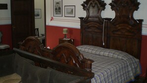 Un dormitorio con cama de madera, una silla de madera, una mesita de noche con lámpara y una pared roja con cuadros enmarcados.