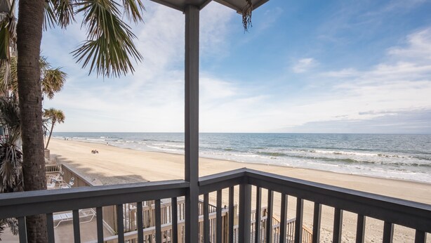 Uitzicht op een strand vanaf een balkon met een leuning, met palmbomen en een heldere lucht.