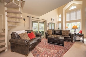 A spacious living room with two leather sofas, a coffee table, a patterned rug, a staircase, and large windows.