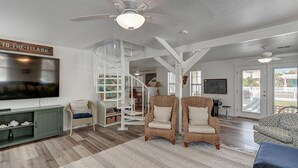 Un vaste salon doté d’un plancher en bois, d’un escalier blanc, de fauteuils en osier, d’un téléviseur et d’une pancarte indiquant « VERS L’ÎLE ».