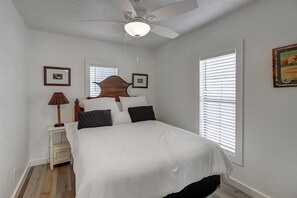 A bedroom with a bed, a nightstand, a lamp, a ceiling fan, and two windows with blinds.