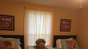 A bedroom with two beds, a nightstand, a window with curtains, and framed pictures on the walls. - Country Living with a Twist (Bolivia)