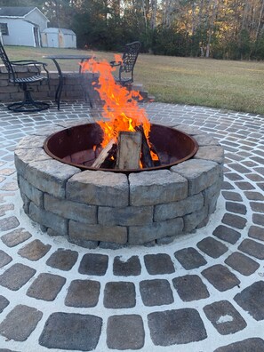 A circular stone fire pit with burning logs, surrounded by a paved patio with a brick pattern.