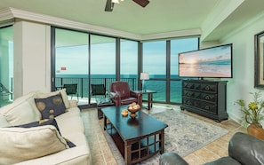 A living room with a sofa, armchair, coffee table, TV, and a balcony with a view of the ocean.