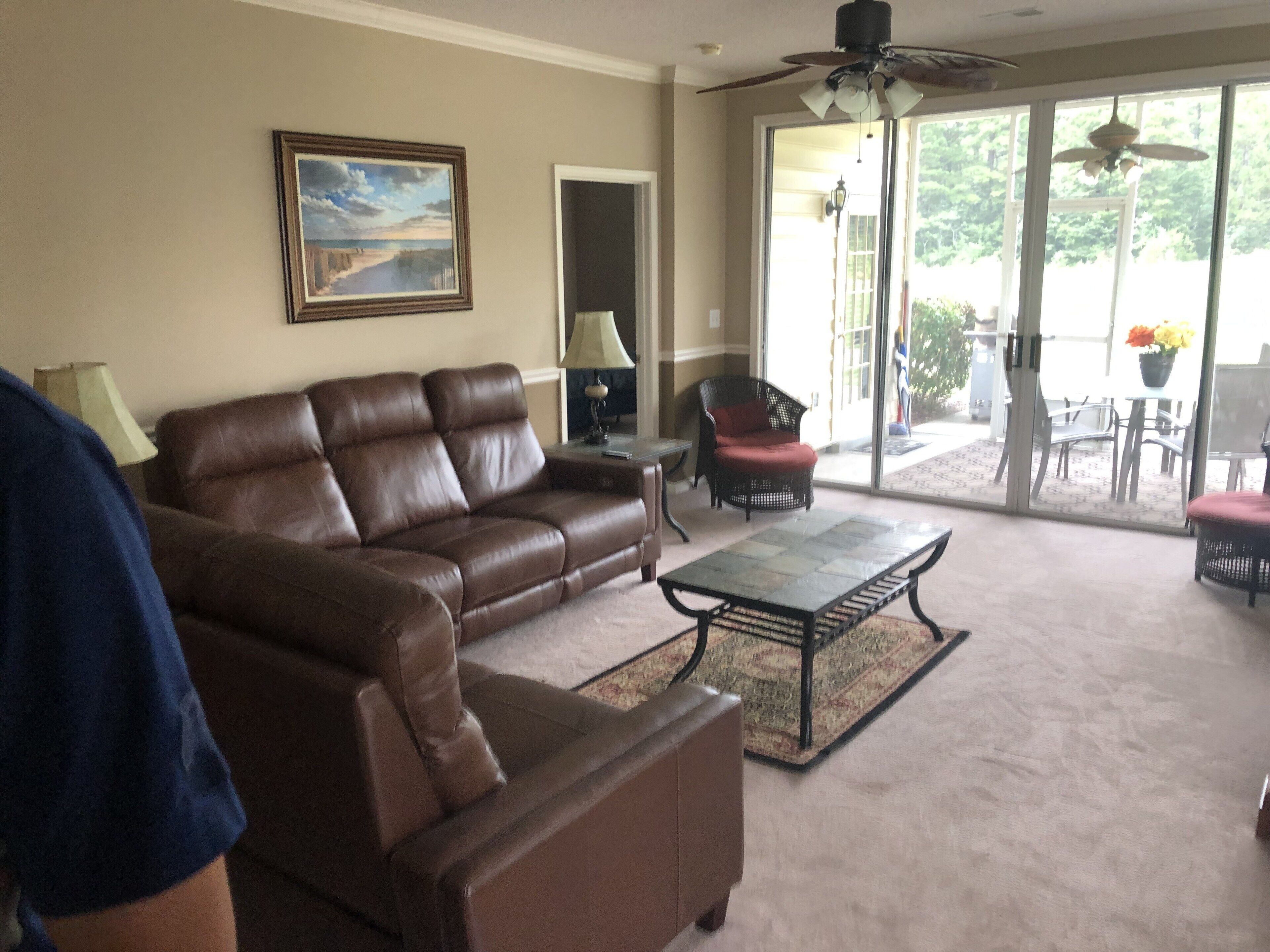A living room with a brown leather sofa, a coffee table, and a chair. There is a painting on the wall and a ceiling fan.
