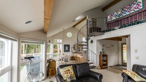 A spacious living room with a black leather sofa, a dining area with a table and chairs, a staircase leading to an upper level, and a large window providing natural light.