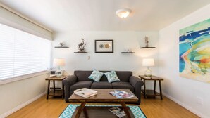 Ein Wohnzimmer mit einer Couch, einem Couchtisch und zwei Beistelltischen mit Lampen. An den Regalen über der Couch befinden sich dekorative Gegenstände und an der Wand ein Gemälde.