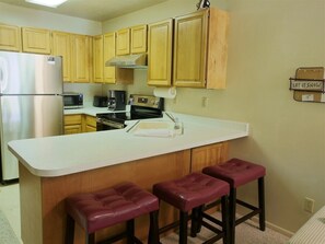 A kitchen with wooden cabinets, a stainless steel refrigerator, a microwave, a stove, and a sink. There are three bar stools at the counter.