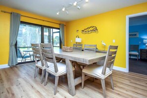 Un coin repas avec une table et des chaises en bois, une fenêtre avec des rideaux et un mur jaune orné d’une fresque représentant des oiseaux.