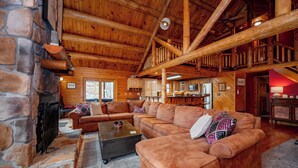 L’intérieur d’une cabane en rondins présente une cheminée en pierre, un grand plafond en bois et un espace de vie chaleureux avec un canapé d’angle et une table basse.