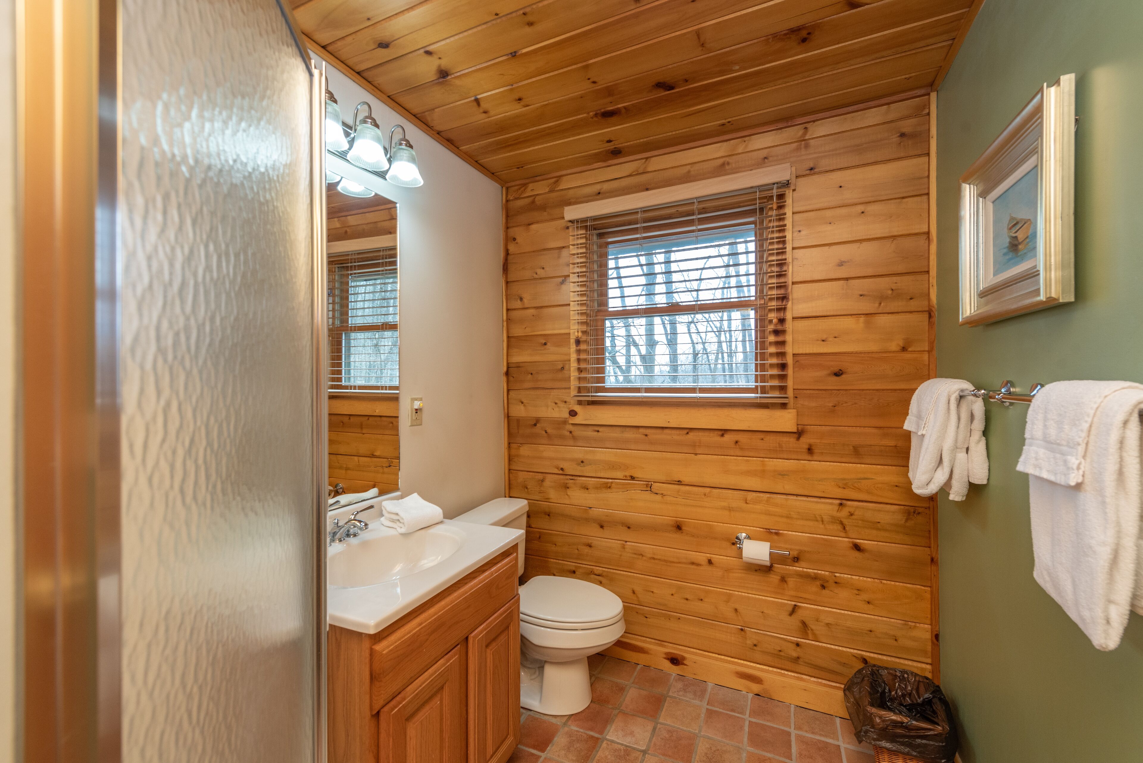 Ein Badezimmer mit Holzwänden, einer weißen Toilette, einem Waschbecken mit Spiegel und einem Fenster mit Jalousien.