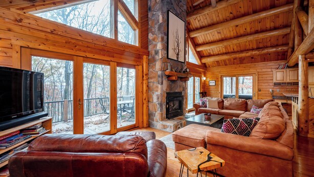 L’intérieur chaleureux d’un chalet avec une cheminée en pierre, des meubles en cuir et de grandes fenêtres donnant sur la forêt.