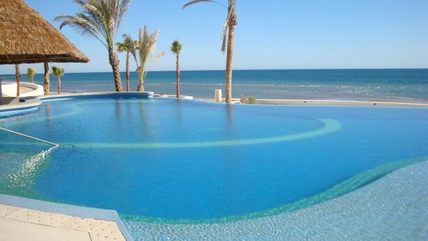 A clear blue infinity pool with a view of the ocean, surrounded by palm trees and a thatched-roof structure. - Tessoro 703 - Award Winning Design at the End of Las Conchas (Puerto Penasco/Rocky Point)