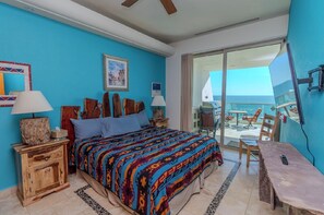 Quarto com uma cama grande, mesa de cabeceira de madeira, uma mesa e vista para o oceano através de uma porta de correr de vidro.