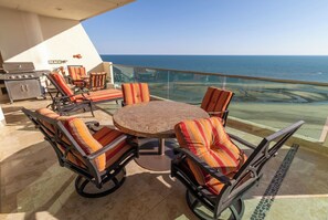 A sunlit outdoor dining area with a round table and striped chairs, overlooking a clear blue sea.