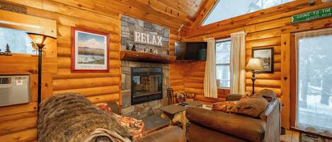 A log cabin interior with a stone fireplace, wooden walls, a television, and a sign reading "GOING-TO-THE-GREAT FALLS".