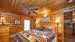 A log cabin-style bedroom with a bed, two wall-mounted lamps, a ceiling fan, and a painting on the wall.