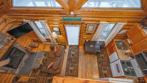 A cabin interior with a fireplace, wooden flooring, a sofa, a dining table, and a kitchen area.