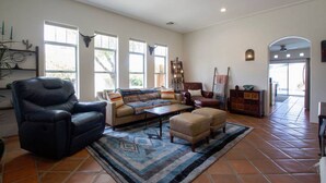 Amplia sala de estar con un sillón de cuero, un sofá y una mesa de centro. Hay ventanas grandes y una puerta que da a otra habitación.