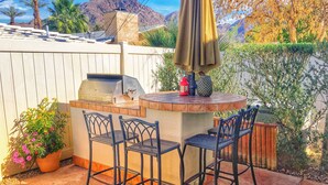 Un patio al aire libre con una parrilla, dos sillas de metal y una mesa con un farol rojo y un jarrón decorativo.