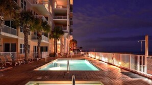 Un área de piscina nocturna con una luna en el cielo, una terraza de madera y un edificio con balcones.