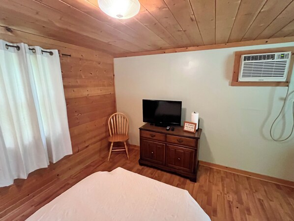 Una habitación con paneles de madera, una cama, un televisor sobre una cómoda, una silla y un aire acondicionado de pared.