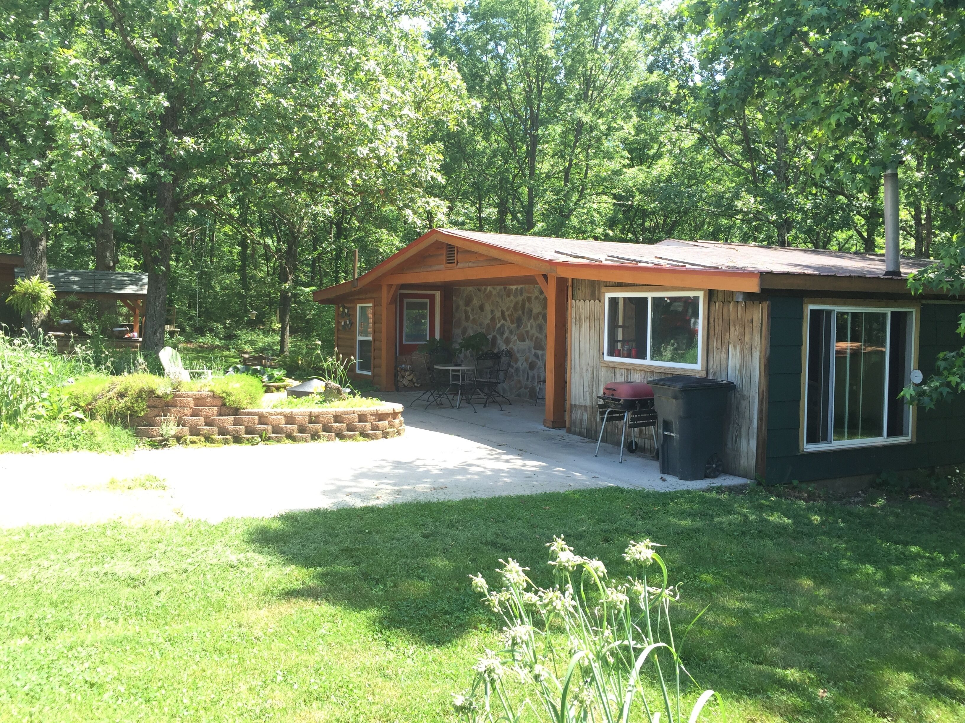 A small wooden cabin with a stone wall, a patio with a table and chairs, and a barbecue grill.