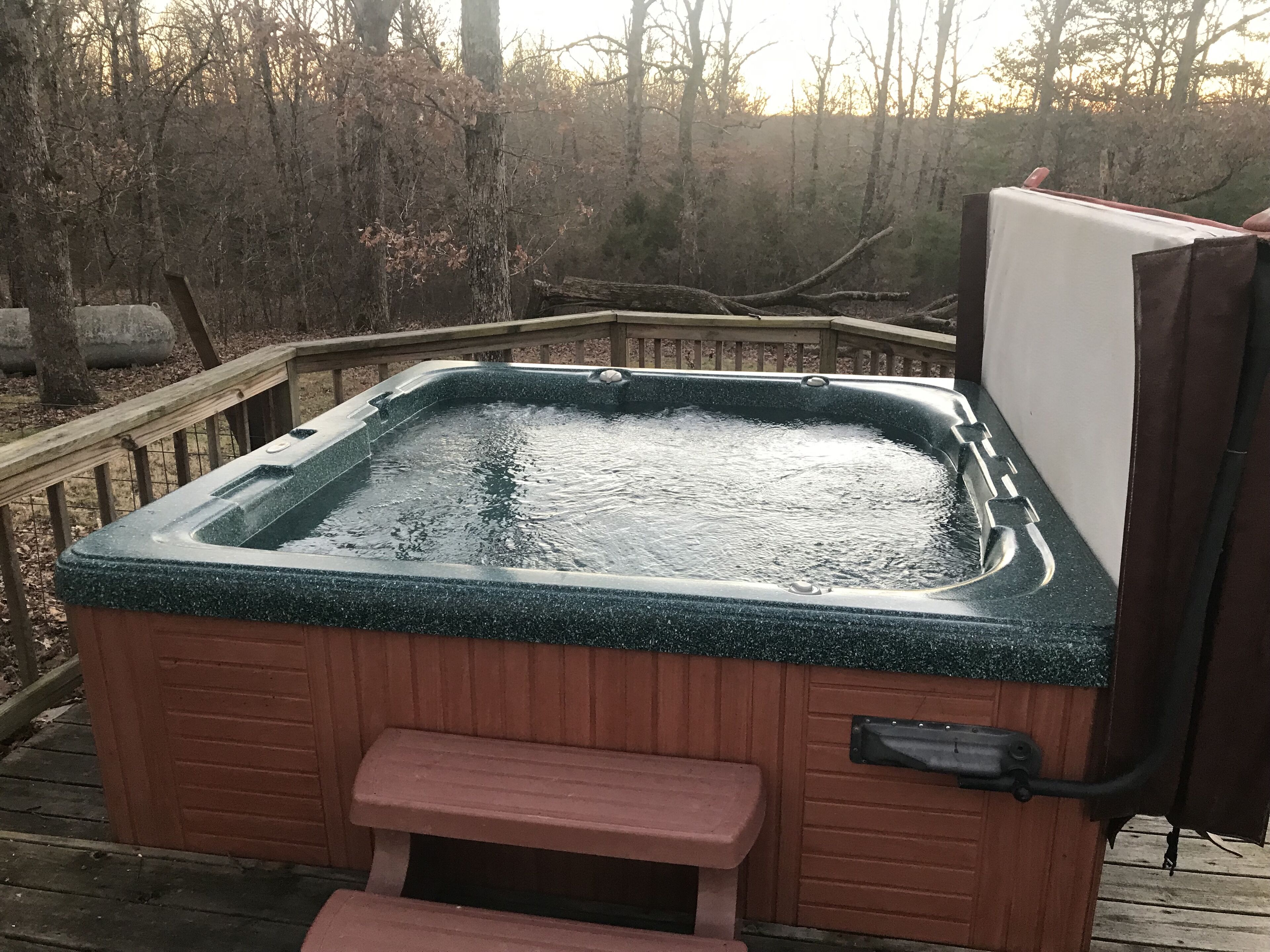 A hot tub with a wooden bench and steps, surrounded by a wooden deck and a forested area.
