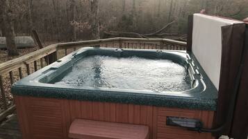 A hot tub with a wooden bench and steps, surrounded by a wooden deck and a forested area.