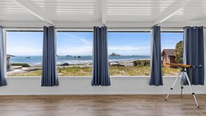 Una habitación con amplios ventanales que dan a una playa, un telescopio sobre un trípode y suelo de madera.