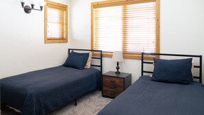 A bedroom with two single beds, a nightstand with a lamp, and two windows with blinds.