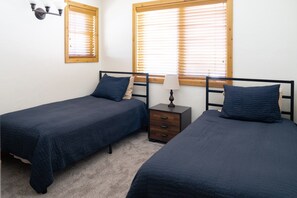 A bedroom with two single beds, a nightstand with a lamp, and two windows with blinds.