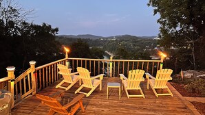 A wooden deck with four Adirondack chairs and a small table, overlooking a river and hills.