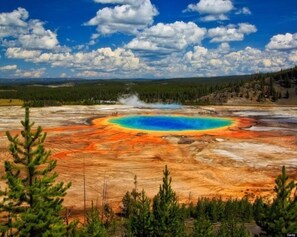 A geothermal hot spring with a vibrant blue center surrounded by orange and red mineral deposits, set against a backdrop of a clear blue sky and forested hills.