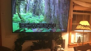 A living room with a television mounted on the wall displaying a forest scene. - 30 Miles from Yellowstone Park! View is Breathtaking! (Emigrant)