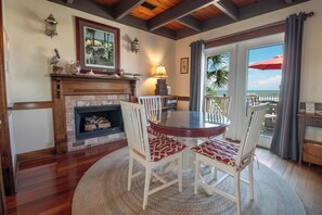 A room with a fireplace, a round table with chairs, a view of the ocean, and a balcony with a red umbrella.