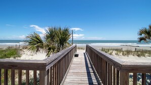 Een houten looppad dat naar een strand leidt, met palmbomen en een helderblauwe lucht.