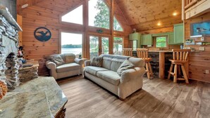 Ein gemütliches Blockhaus-Interieur mit Kamin aus Stein, einer Couch, einer Essecke mit Holzsesseln und großen Fenstern mit Blick ins Freie.