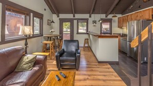Amplia sala de estar con un sofá de cuero, una mesa de centro de madera y un sillón negro. Se observa una cocina con armarios de madera y electrodomésticos de acero inoxidable al fondo.