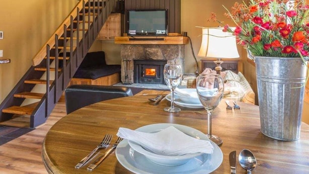 Un comedor con una mesa de madera preparada para dos personas, una chimenea y una escalera.